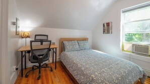 Ein Schlafzimmer mit einem Bett, einem Schreibtisch mit Stuhl, einer Lampe und einem Fenster mit Klimaanlage.