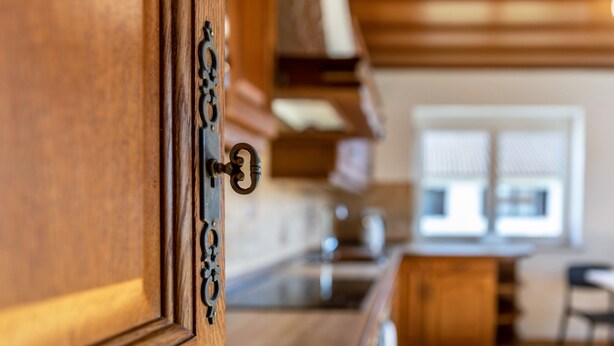 A wooden cabinet with a keyhole, a kitchen area with a countertop, and a window with blinds. - Attic Apartment with Air Conditioning in Freilassing (Freilassing)