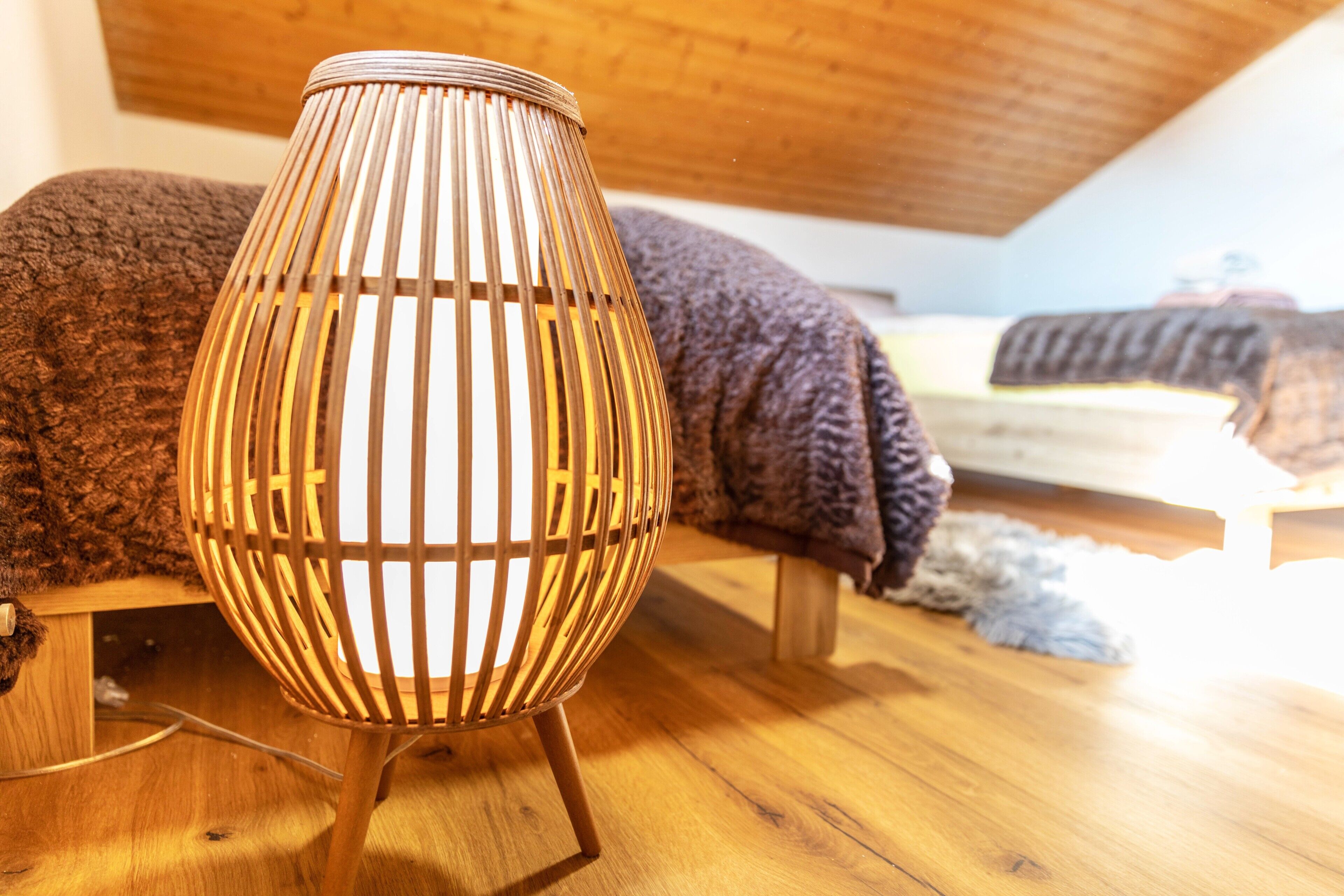 Ein Zimmer mit Holzboden, einem Bett mit dunkler Decke und einer Holzlampe mit gerippter Struktur.