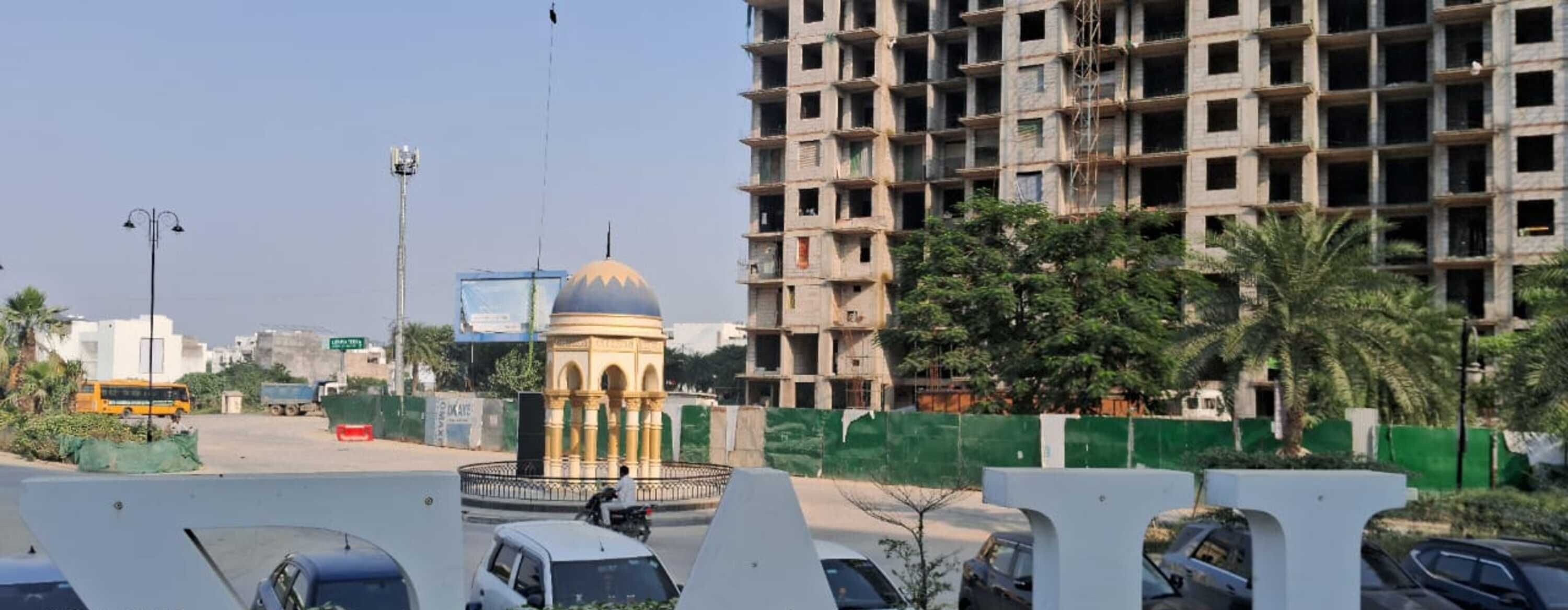 A view of a cityscape with a large, unfinished building, a roundabout, and parked cars.