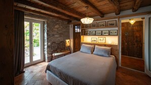 Une chambre avec un mur en pierre, des poutres en bois au plafond, un lit recouvert d’une courtepointe, une chaise et une porte avec un panneau en bois.