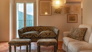 Un salon avec un canapé à motifs, un fauteuil blanc et une petite table assortie d’un pouf. Des œuvres d’art encadrées ornent le mur, et un balcon offre une vue sur les collines.