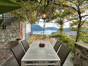 Terraço com mesa e cadeiras, um muro de pedra, luminária pendente e vista para um lago e montanhas.