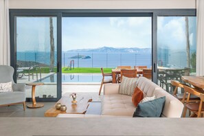 Een moderne woonkamer met een grote schuifpui naar een zwembadgedeelte, een bank met kussens, een salontafel en een eettafel met stoelen.