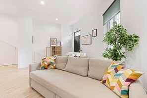 A modern living room with a beige sofa, colorful chevron pillows, a potted plant, and framed artwork on the wall.