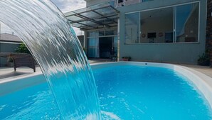 Ein Pool mit Wasserfall, eine überdachte Terrasse und ein modernes Haus mit großen Fenstern.