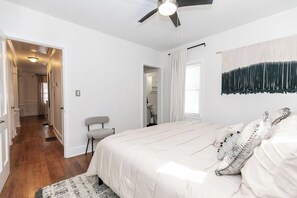 Une chambre à coucher avec un lit, un ventilateur de plafond, une fenêtre avec des rideaux et un couloir menant à une autre pièce.