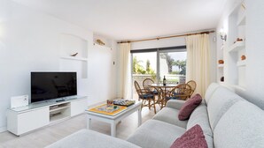 Un salon moderne avec un téléviseur à écran plat, une table basse sur laquelle est posé un jeu de société, un canapé gris et un coin repas avec des chaises et une table en osier.