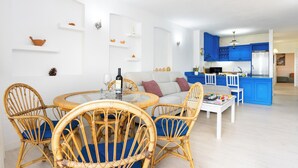 Un salon moderne avec une table ronde en verre, des fauteuils en osier et un canapé. On aperçoit, au fond, une cuisine avec des meubles bleus et un réfrigérateur en acier inoxydable.