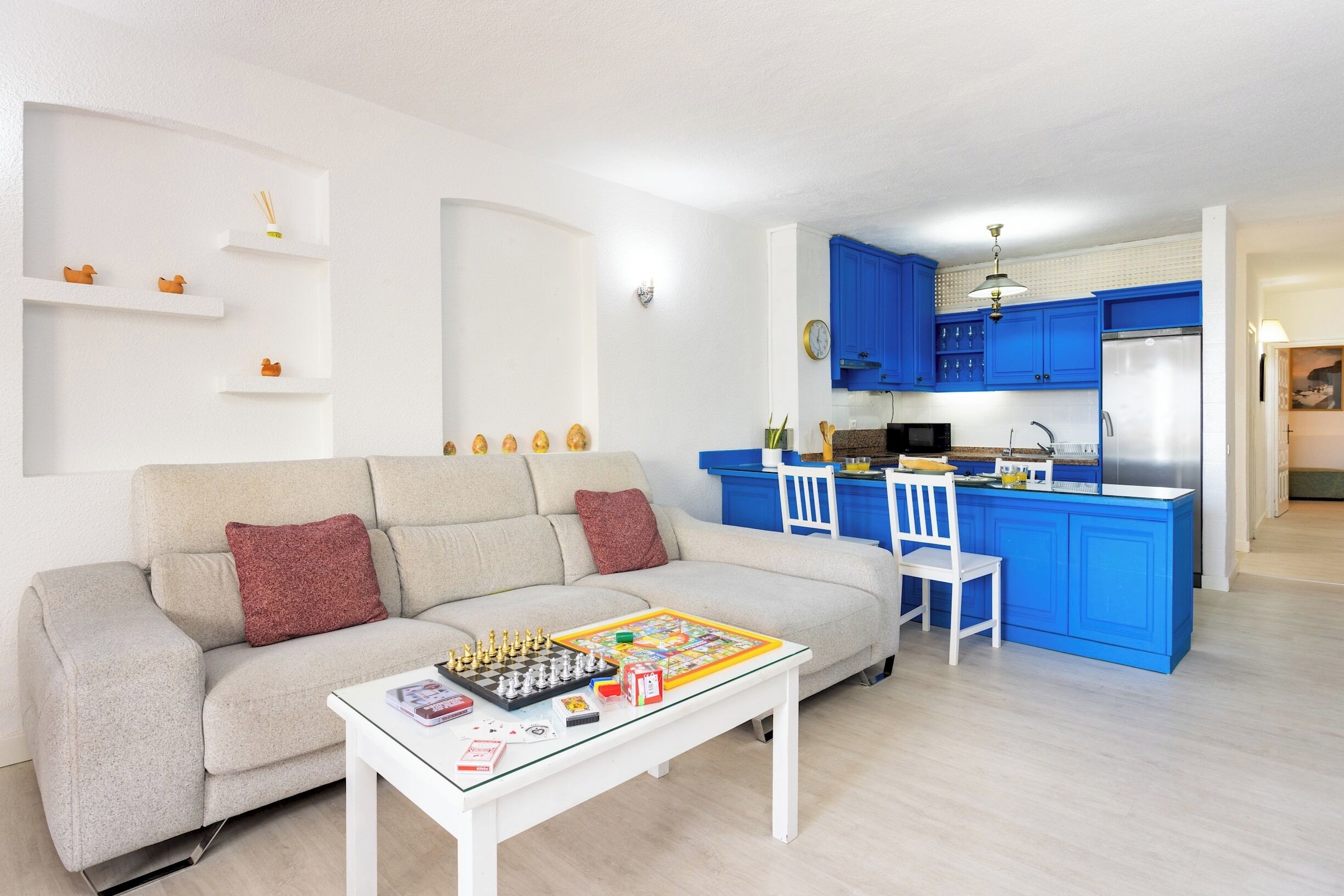 A living room with a sofa, coffee table, and a chessboard. There is a kitchen area with blue cabinets and a refrigerator.