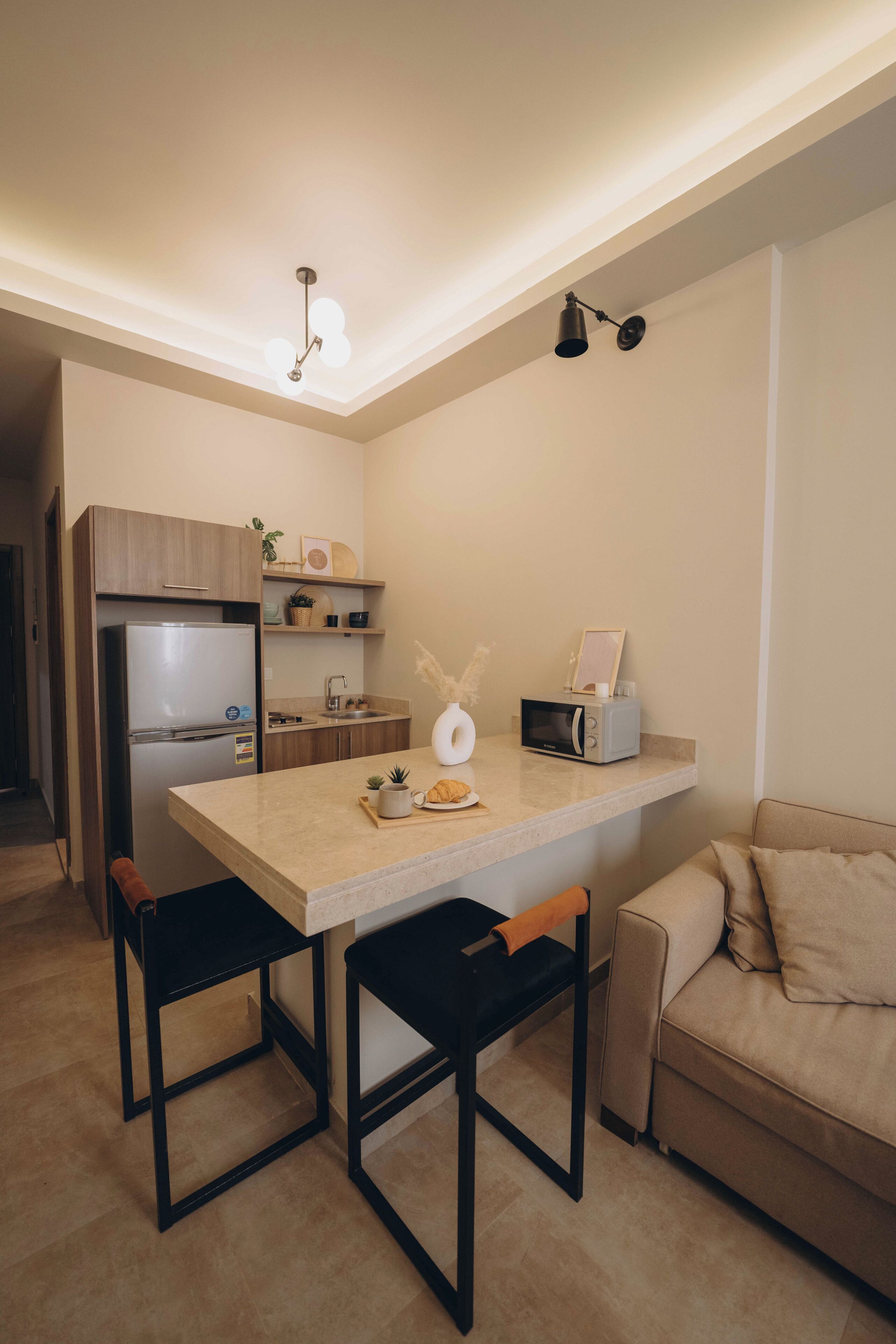 Una cocina compacta con refrigerador, microondas y una pequeña encimera. En la zona de estar contigua hay dos sillas y un sofá.