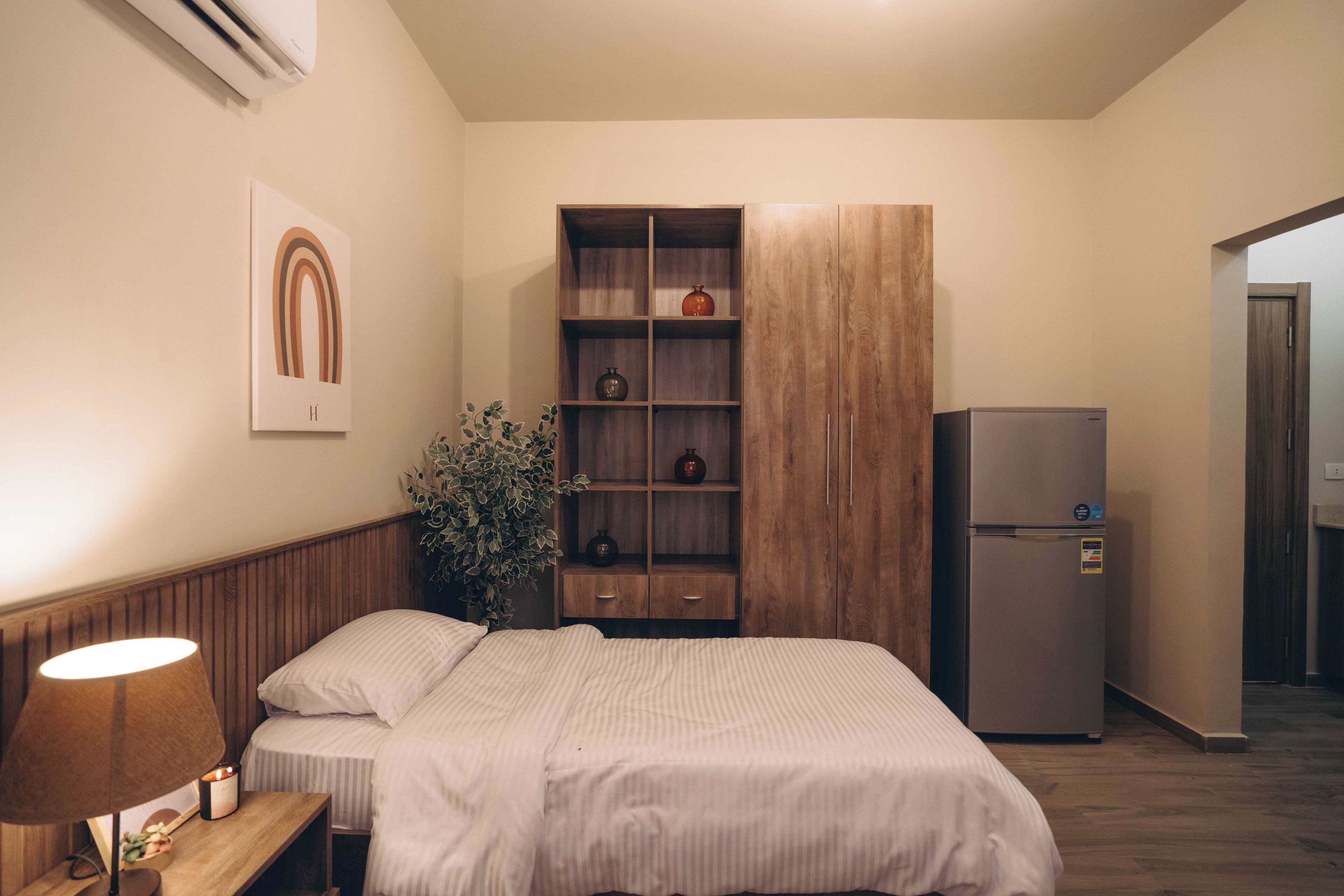 A bedroom with a bed, bedside table with a lamp, a wooden wardrobe, and a refrigerator.