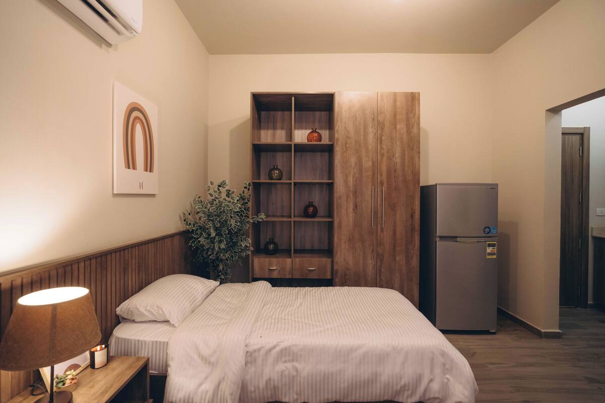 A bedroom with a bed, bedside table with a lamp, a wooden wardrobe, and a refrigerator.