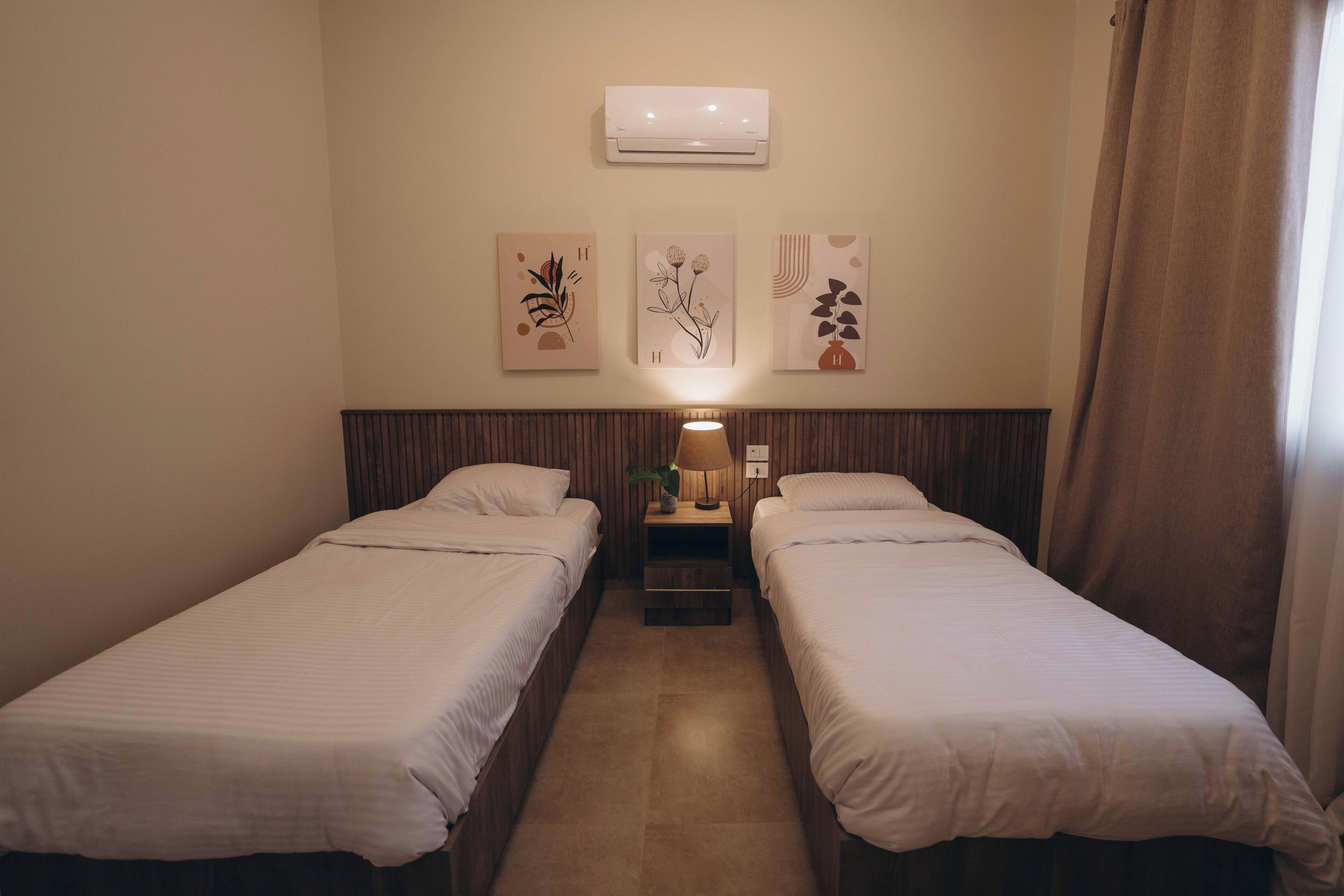 Une chambre d’hôtel avec deux lits simples, une table de chevet avec une lampe, un climatiseur fixé au mur et trois œuvres d’art encadrées.