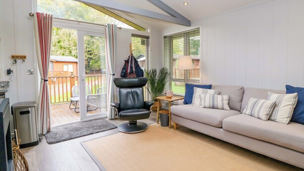 A modern living room with a sofa, a chair, a small table, and a lamp. There is a sliding glass door leading to a patio area. - 5 Waterside Wood (Windermere)