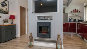 A modern living room with a flat-screen TV mounted above a fireplace, wooden flooring, and a kitchen area in the background. - 5 Waterside Wood (Windermere)