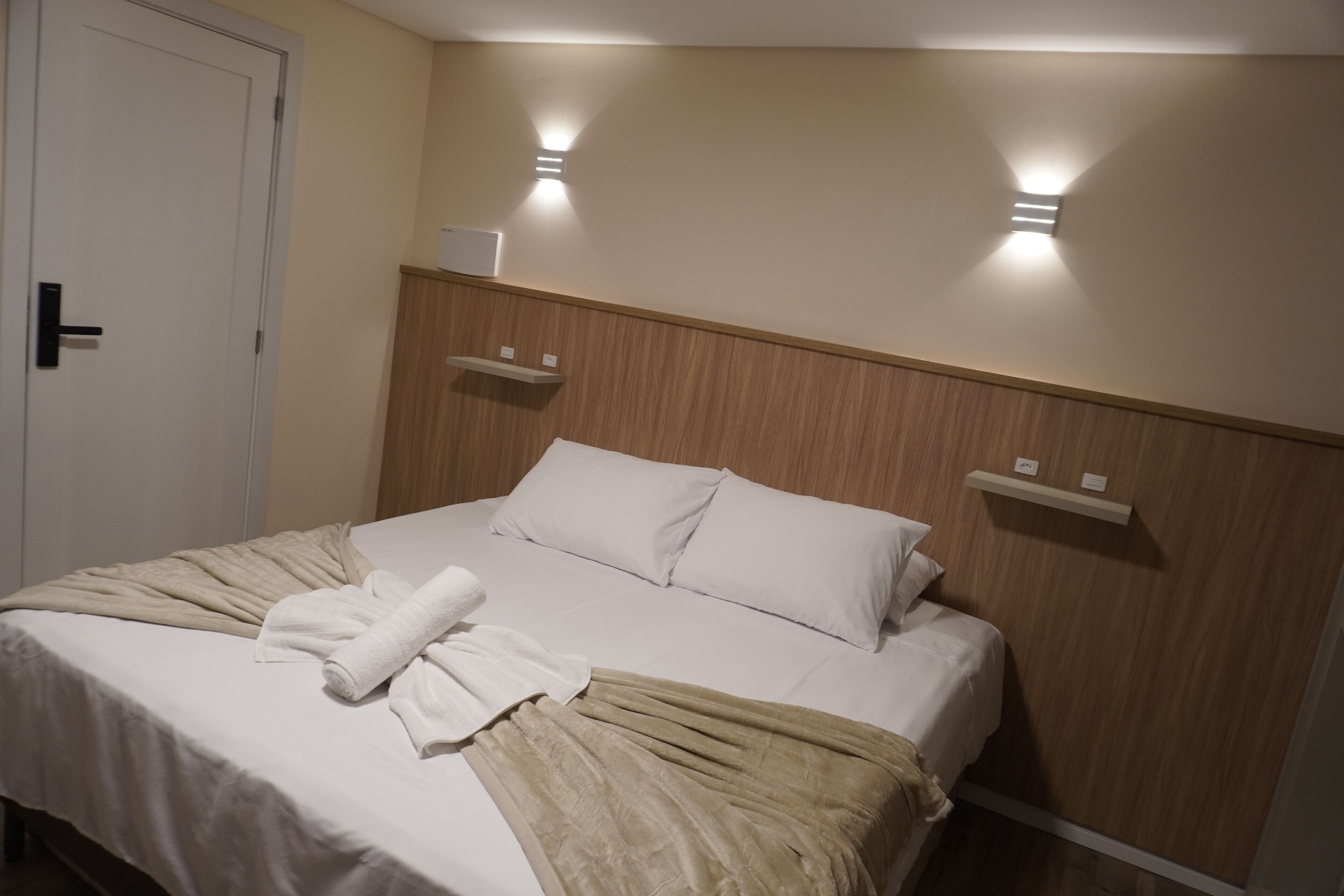 A neatly made bed with white linens, a beige throw blanket, and a folded towel. There are two wall-mounted light fixtures on either side of the headboard.