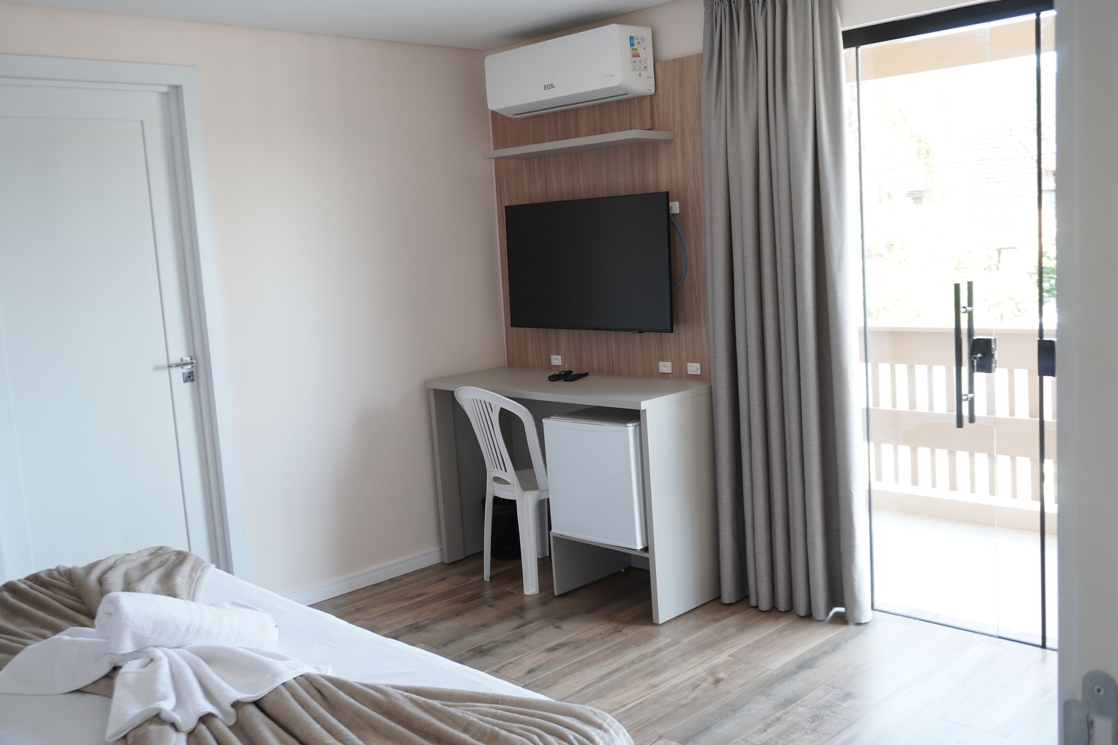 A hotel room with a bed, a desk with a chair, a flat-screen TV, and an air conditioning unit.