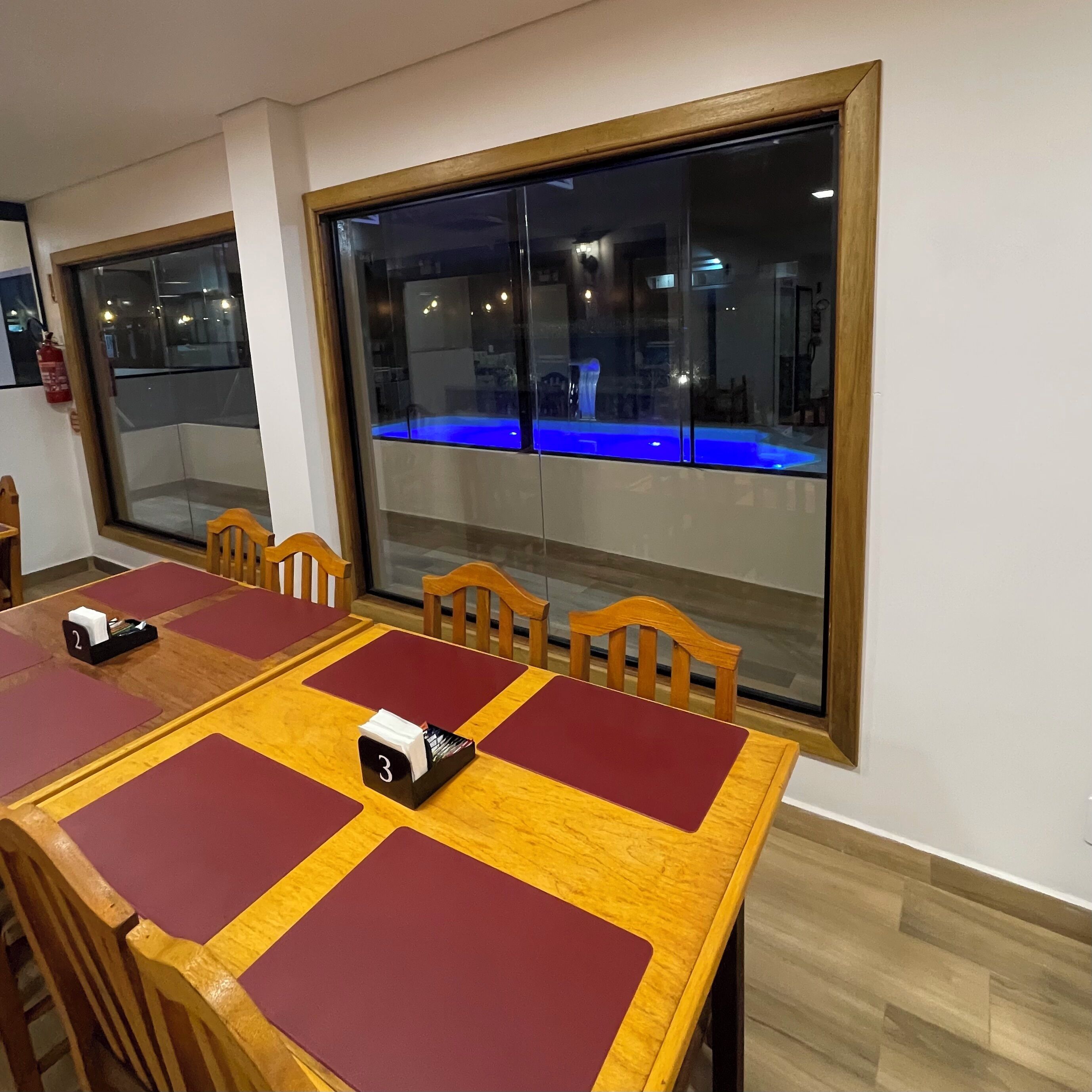 A dining area with wooden tables and chairs, a fire extinguisher, and a view of a blue-lit pool through large windows.