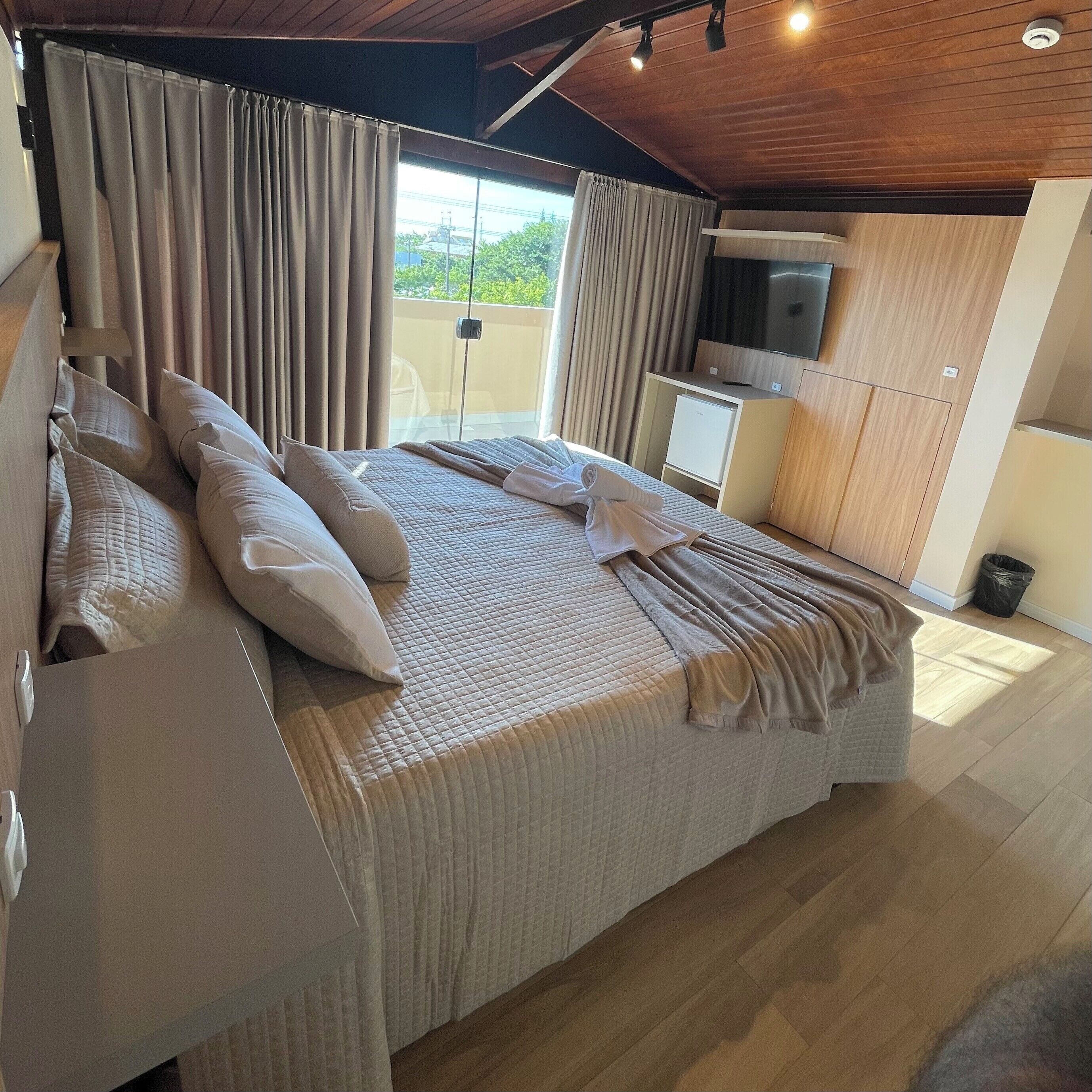 A modern bedroom with a large bed, curtains, a TV, and a small kitchenette.