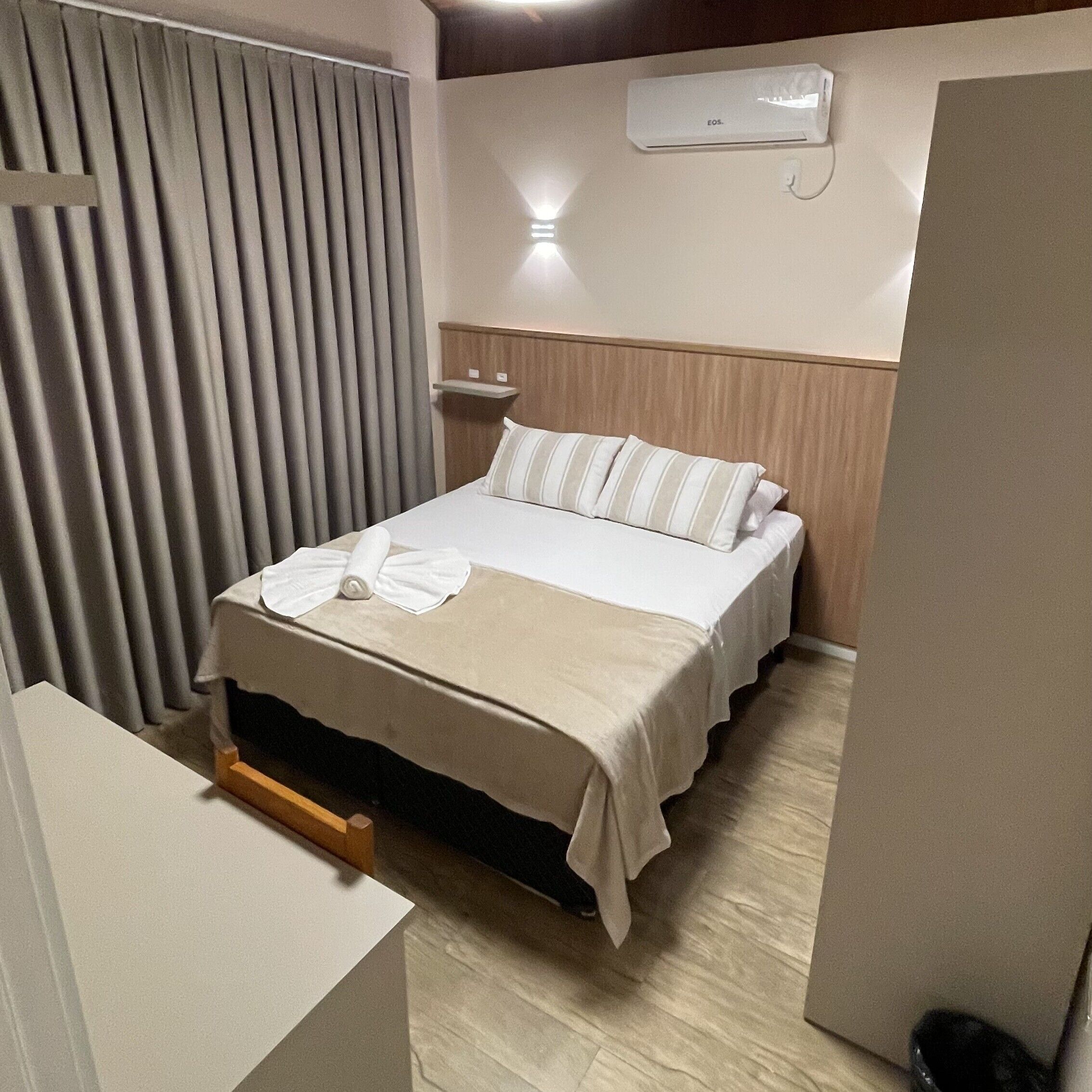 A hotel room with a bed, a bedside table, a wall-mounted air conditioner, and a curtain.