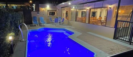 Una piscina con tonalidades azules, rodeada por una terraza con sillas y una zona de estar cubierta.
