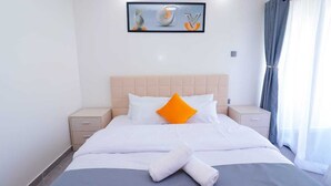 A neatly made bed with white linens and an orange pillow, a framed artwork above it, and a window with blue curtains.