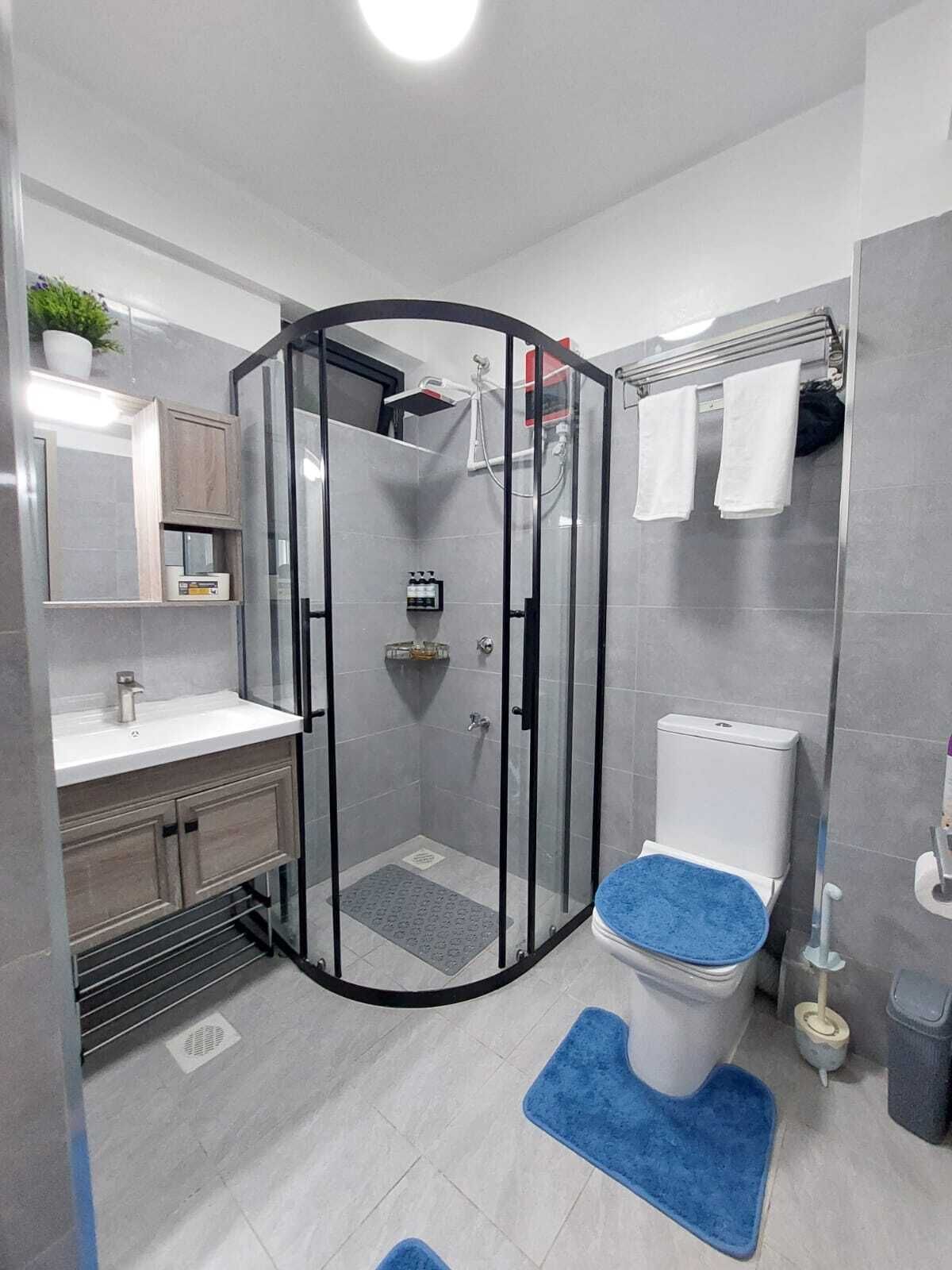 Une salle de bain moderne avec une douche à parois de verre, des toilettes avec un abattant bleu, un lavabo avec un miroir au-dessus et un porte-serviettes avec des serviettes.