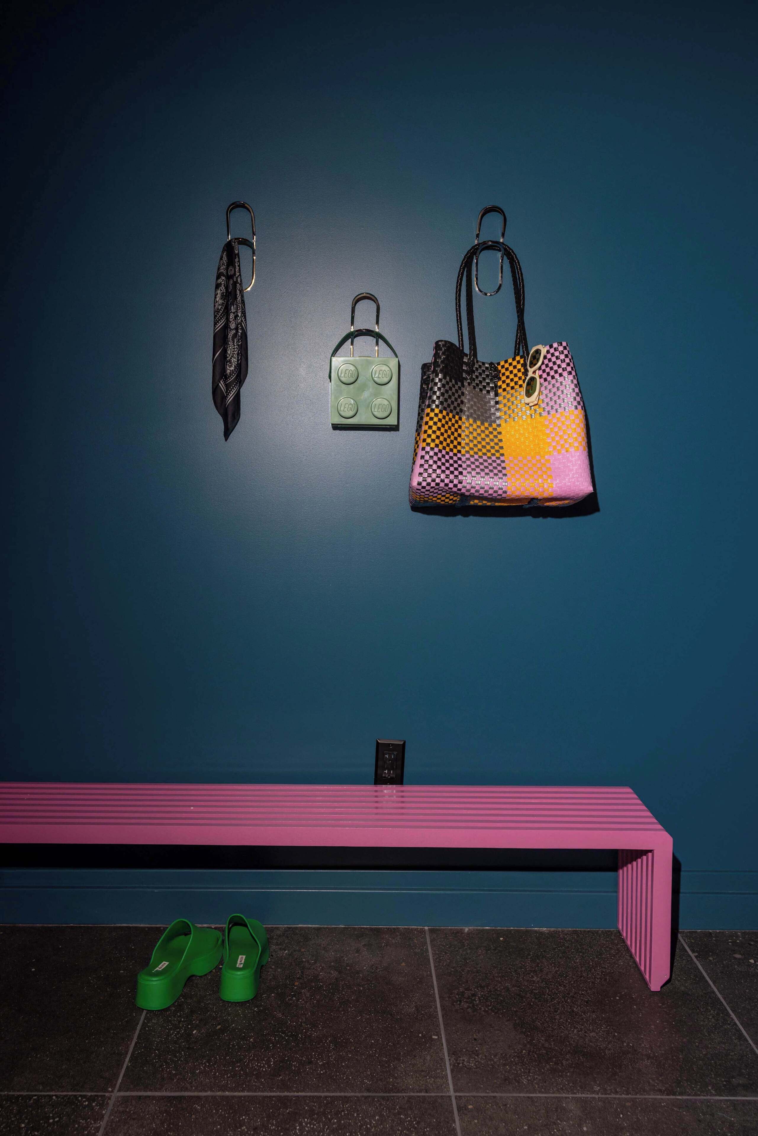 A bench with a pair of green slippers beside it, a wall-mounted bag rack with three bags, and a power outlet on the wall.