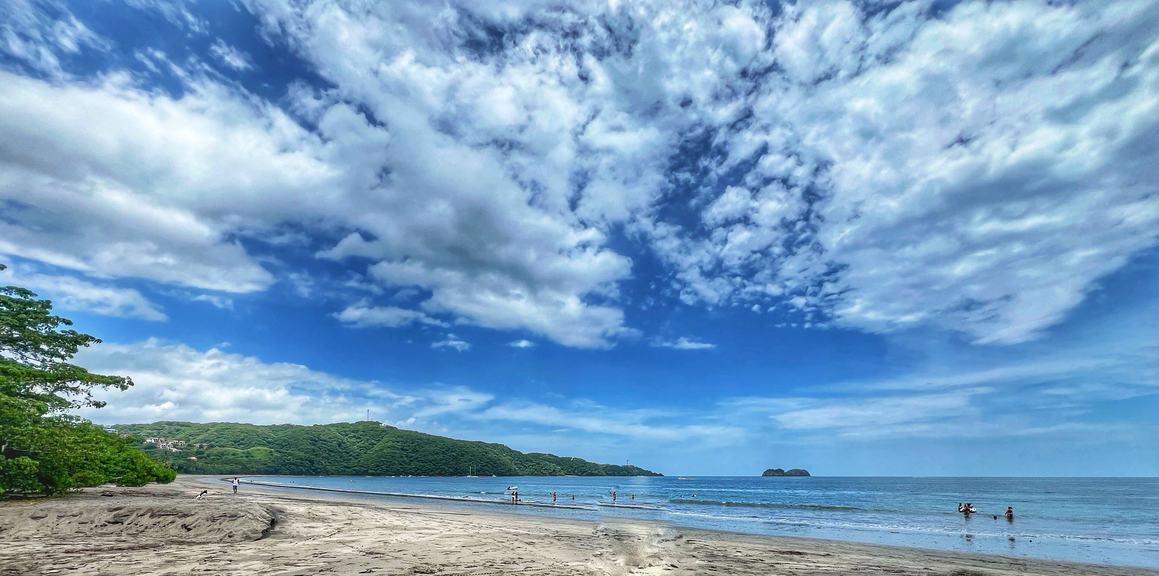 Una playa con cielo despejado y mar tranquilo, rodeada de vegetación y colinas a lo lejos.