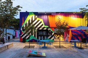 An outdoor seating area with tables and chairs, a mural on the building, and a small tree.