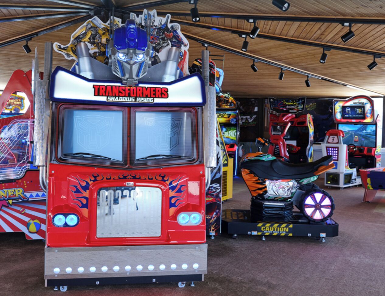 A Transformers-themed arcade machine with a red and silver design, surrounded by other arcade games.