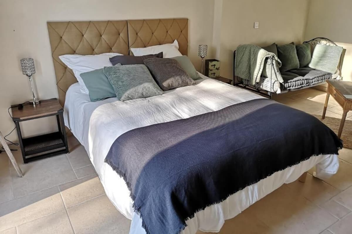 A neatly made bed with a tufted headboard, a bedside table with a lamp, and a chair with a blanket.
