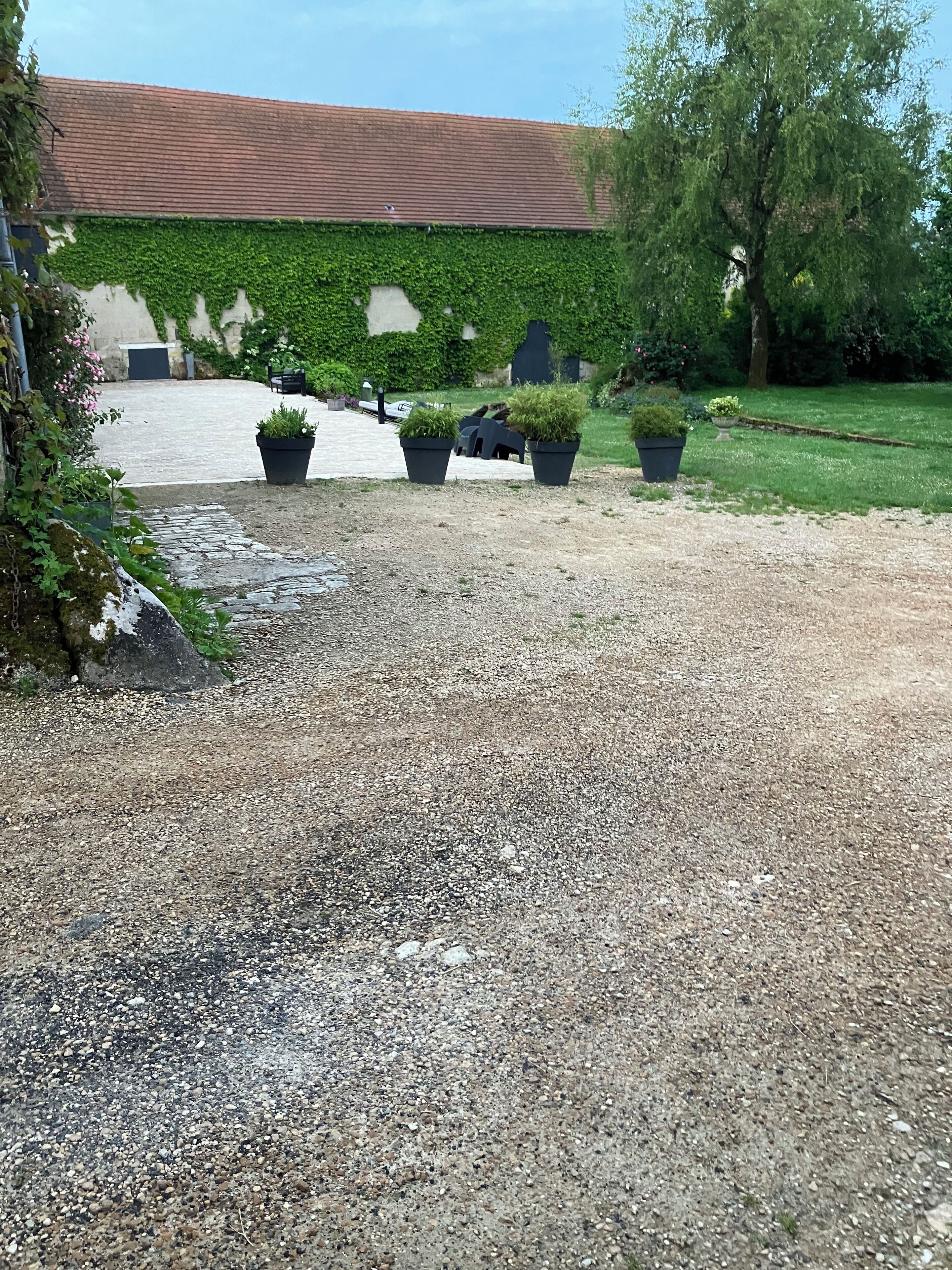 A gravel driveway leading to a building with a red roof, surrounded by potted plants and greenery.
