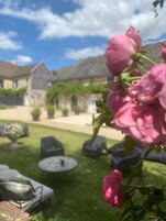 Um jardim com um arbusto de rosas cor-de-rosa em primeiro plano, um edifício de pedra ao fundo e área de estar externa com mesas e cadeiras redondas.