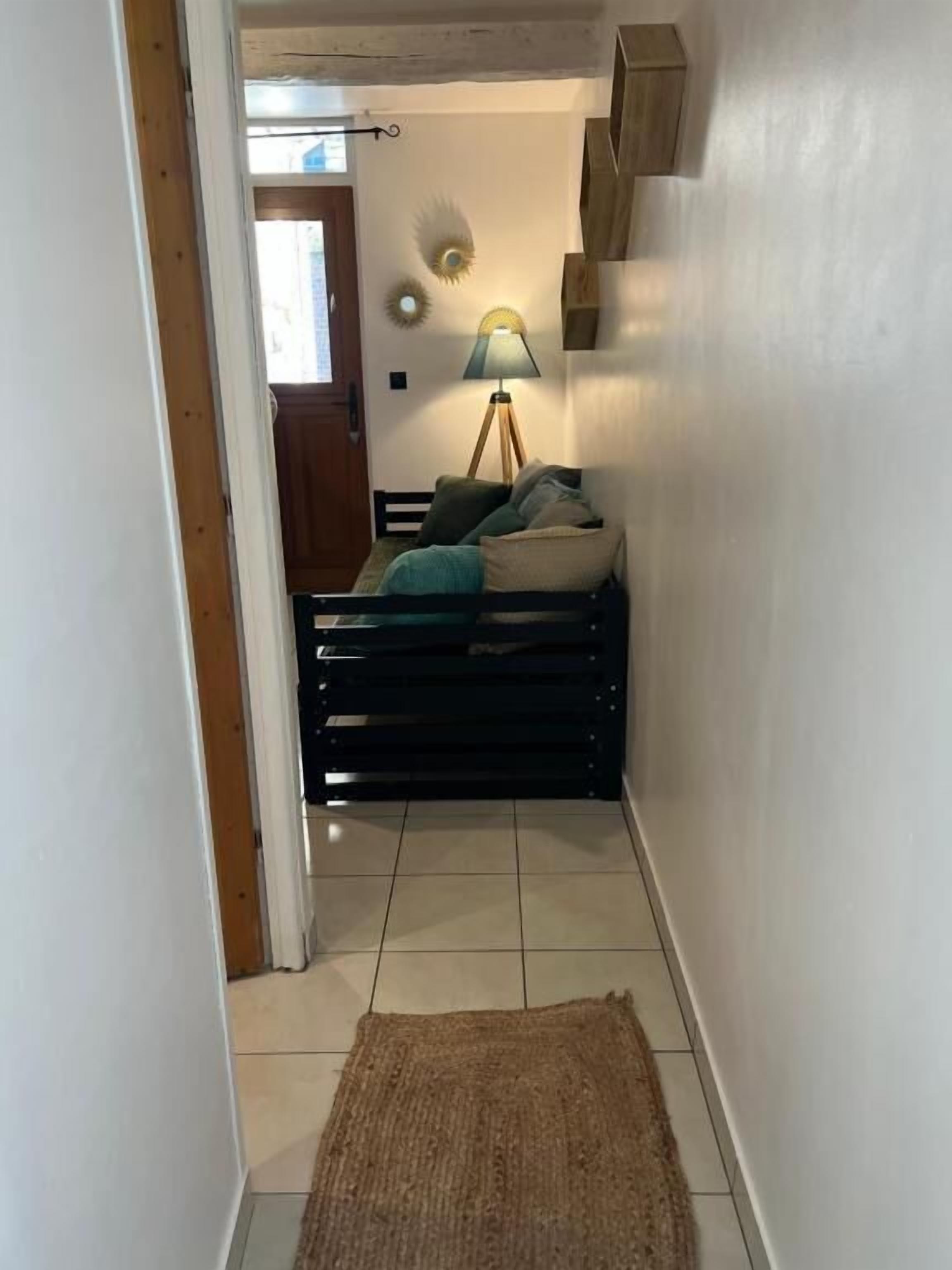 A narrow hallway with a wooden door at the end, a small rug on the floor, and a bed with pillows and a lamp in the background.