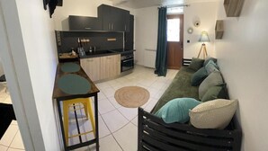 A compact living space with a kitchenette, a sofa with cushions, a small round table, and a lamp. - La ferme de Valeuse (Morienval)