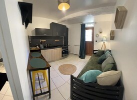 Um espaço de estar compacto com cozinha compacta, um sofá com almofadas, uma pequena mesa redonda e uma luminária.