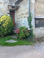 Um edifício de pedra com porta de madeira, caixa de correio e um pequeno jardim com arbustos verdes e flores cor-de-rosa.