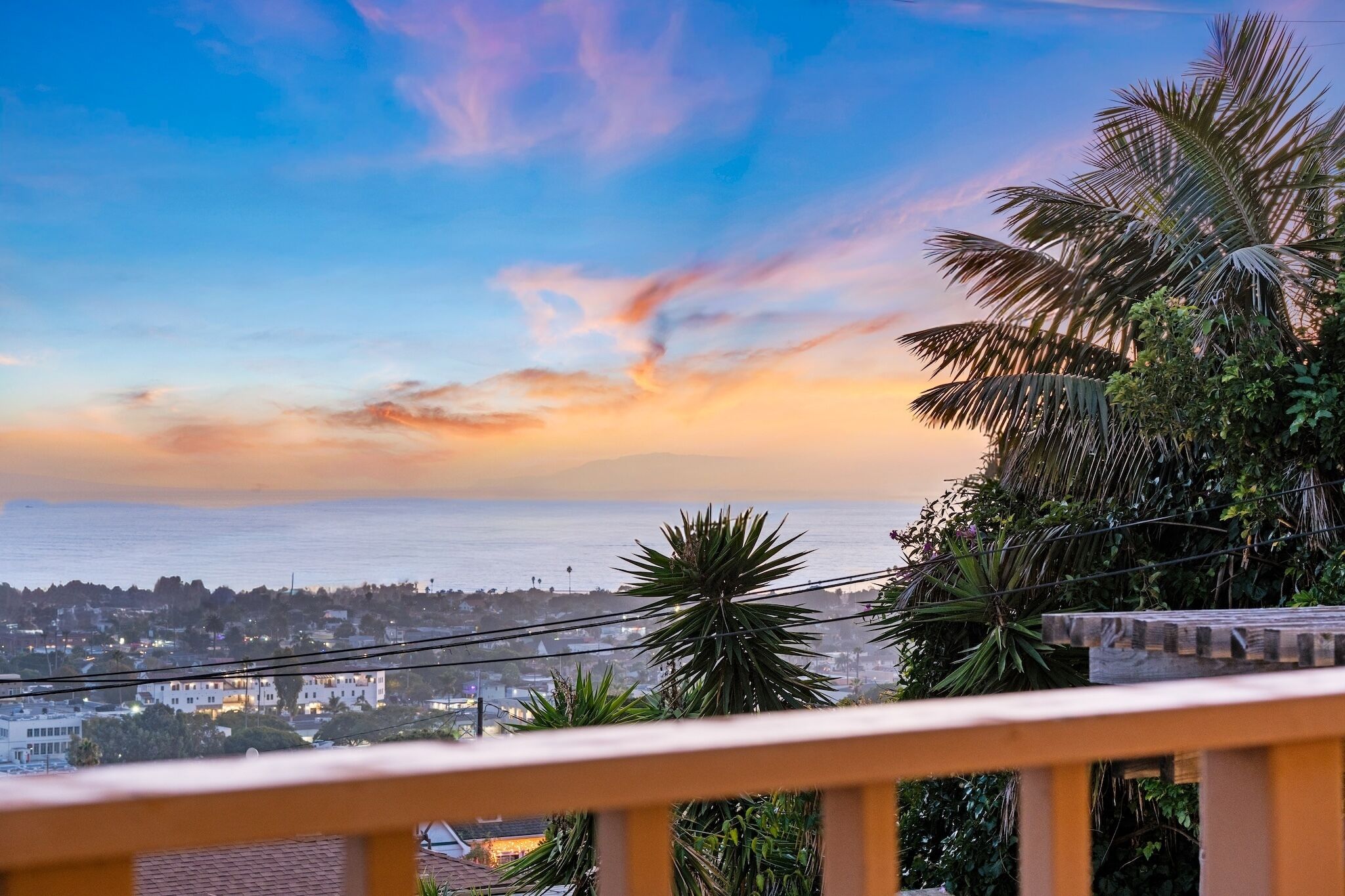 A coastal cityscape with a sunset, palm trees, and a balcony railing.