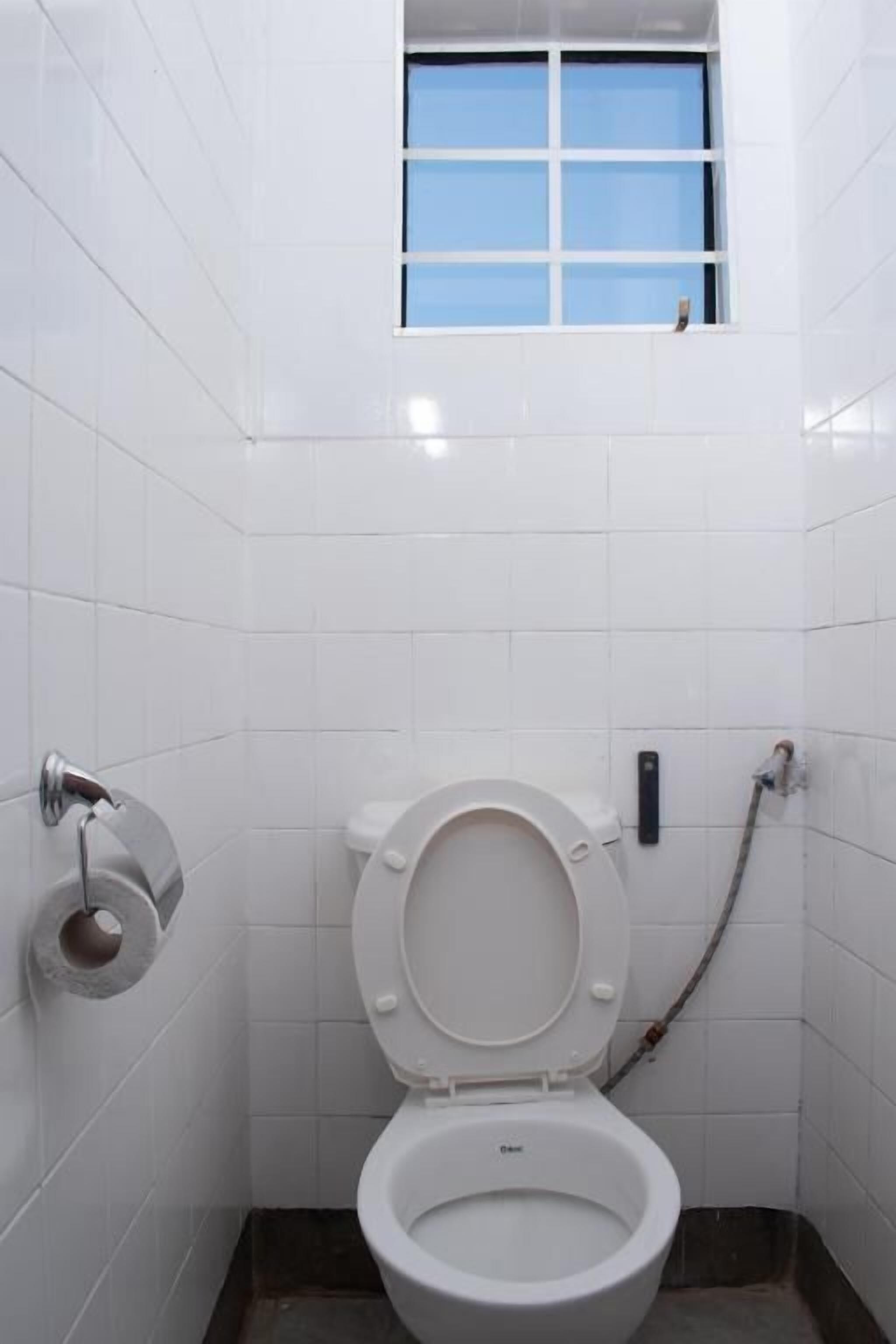 Une petite salle de bain aux murs recouverts de carreaux blancs, avec des toilettes, une fenêtre et une tuyauterie apparente.