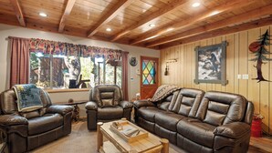 A cozy living room with leather sofas, a wooden coffee table, and a large window with curtains. - All in Cabin (Big Bear Lake)