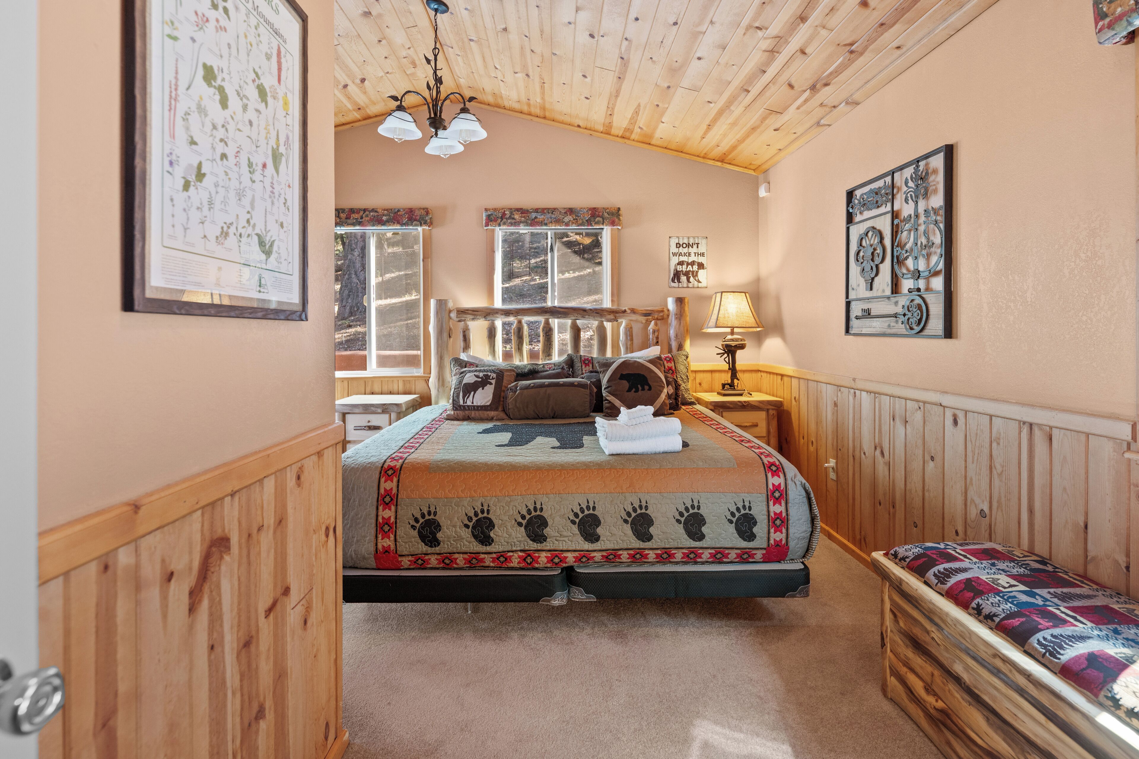 A cozy bedroom with a wooden ceiling, a large bed with a patterned bedspread, a bench, and a wall-mounted lamp.