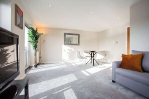 Ein modernes Wohnzimmer mit einem grauen Sofa, einem Fernseher, einem Esstisch mit Stühlen und einer großen Pflanze.