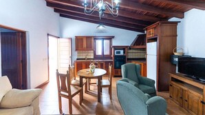 Un salon chaleureux avec un plafond en bois, un coin repas avec une table et des chaises, une cuisine avec des meubles et des appareils électroménagers en bois, et un téléviseur posé sur un meuble en bois.
