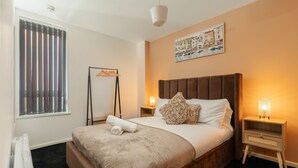 A neatly made bed with a brown headboard, a bedside table with a lamp, and a large window with blinds. - Spacious City Apartment| 2 bd/2 bth| Free Parking (Merseyside)