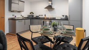 A modern kitchen with a dining area, featuring a glass table, black chairs, and a yellow trash bin. - Spacious City Apartment| 2 bd/2 bth| Free Parking (Merseyside)