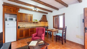 Cocina compacta con armarios de madera, microondas y lavamanos. Hay una zona de comedor con una silla roja y una mesa, y una ventana con persianas.
