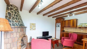 Una sala de estar con chimenea de piedra, vigas de madera, televisor, un sillón rosa y un área de cocina con armarios de madera.