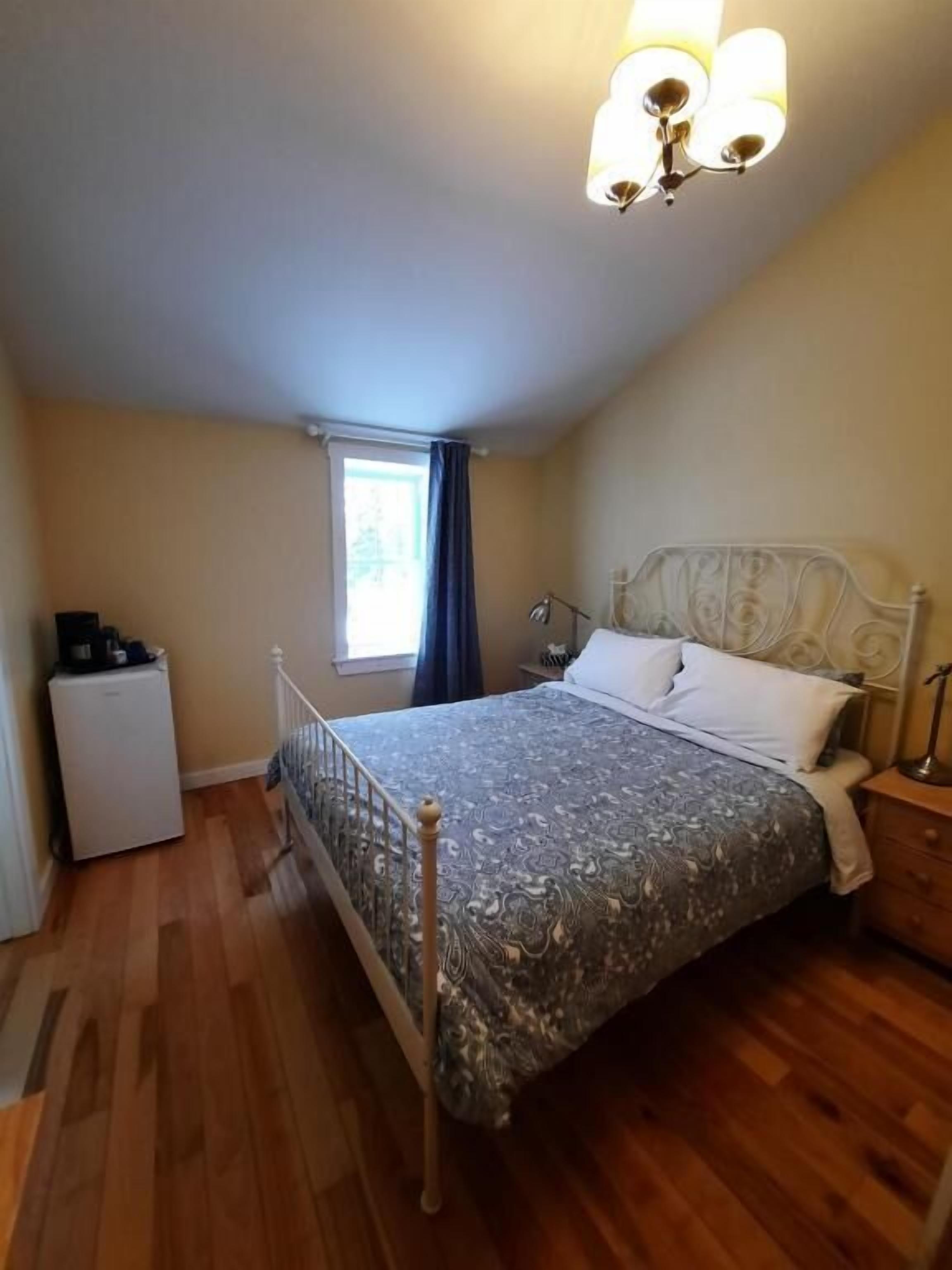 A bedroom with a bed, a nightstand, a lamp, a window, and a small refrigerator.